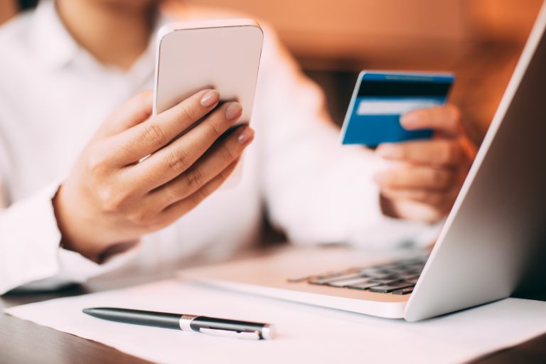 Close-up of female hand holding smartphone. Young businesswoman sitting at laptop in office holding credit card and entering data on her mobile phone
